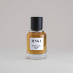 HNKI Hinoki 50 mL botanical perfume in a minimalist glass bottle with a matte black cap and a dark golden hue, set against a pale gray backdrop.