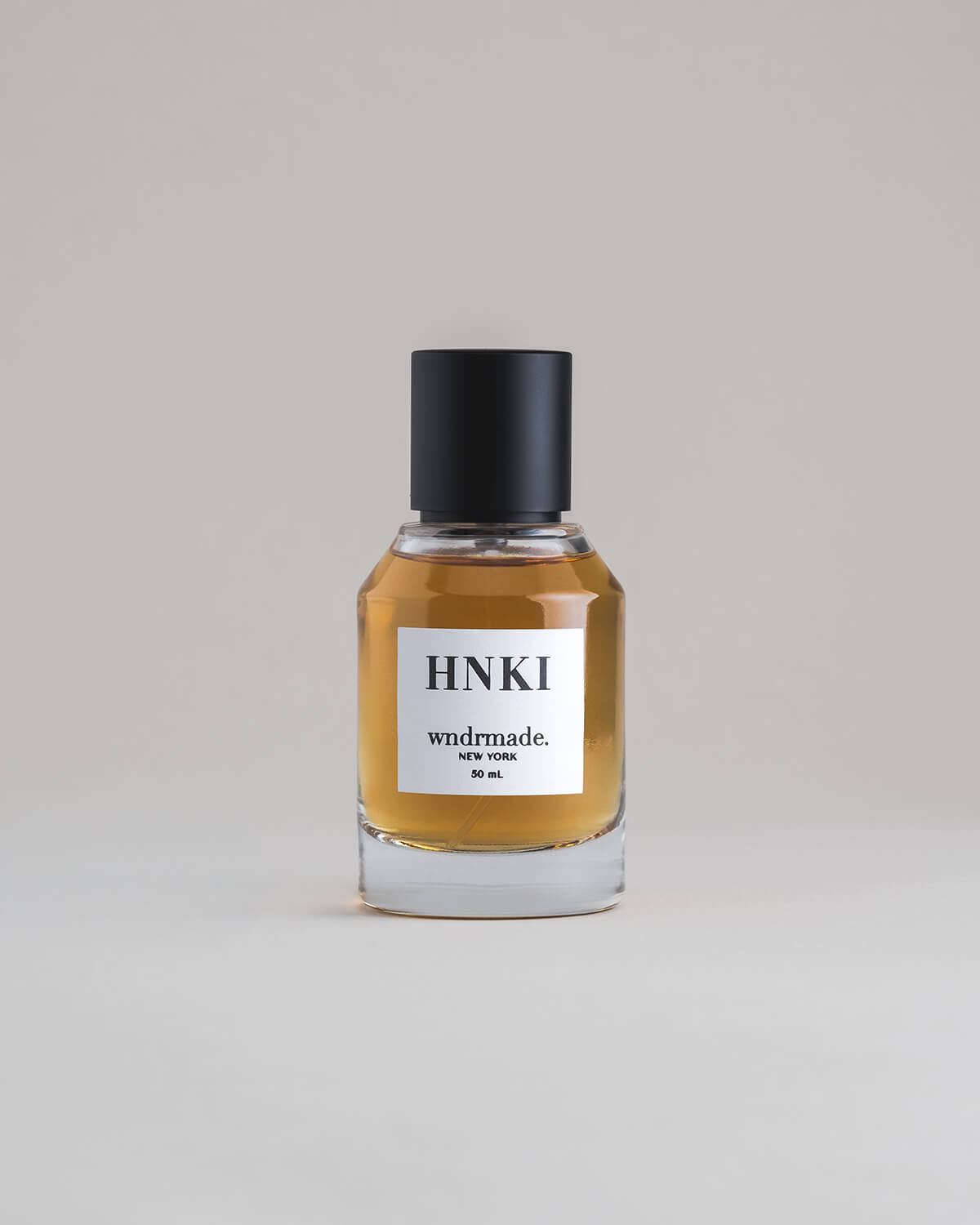 HNKI Hinoki 50 mL botanical perfume in a minimalist glass bottle with a matte black cap and a dark golden hue, set against a pale gray backdrop.