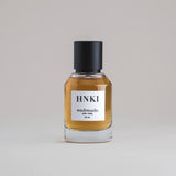 HNKI Hinoki 50 mL botanical perfume in a minimalist glass bottle with a matte black cap and a dark golden hue, set against a pale gray backdrop.