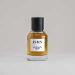JSMN Jasmine 50 mL botanical perfume in a minimalist glass bottle with a matte black cap and a dark golden hue, set against a pale gray backdrop.