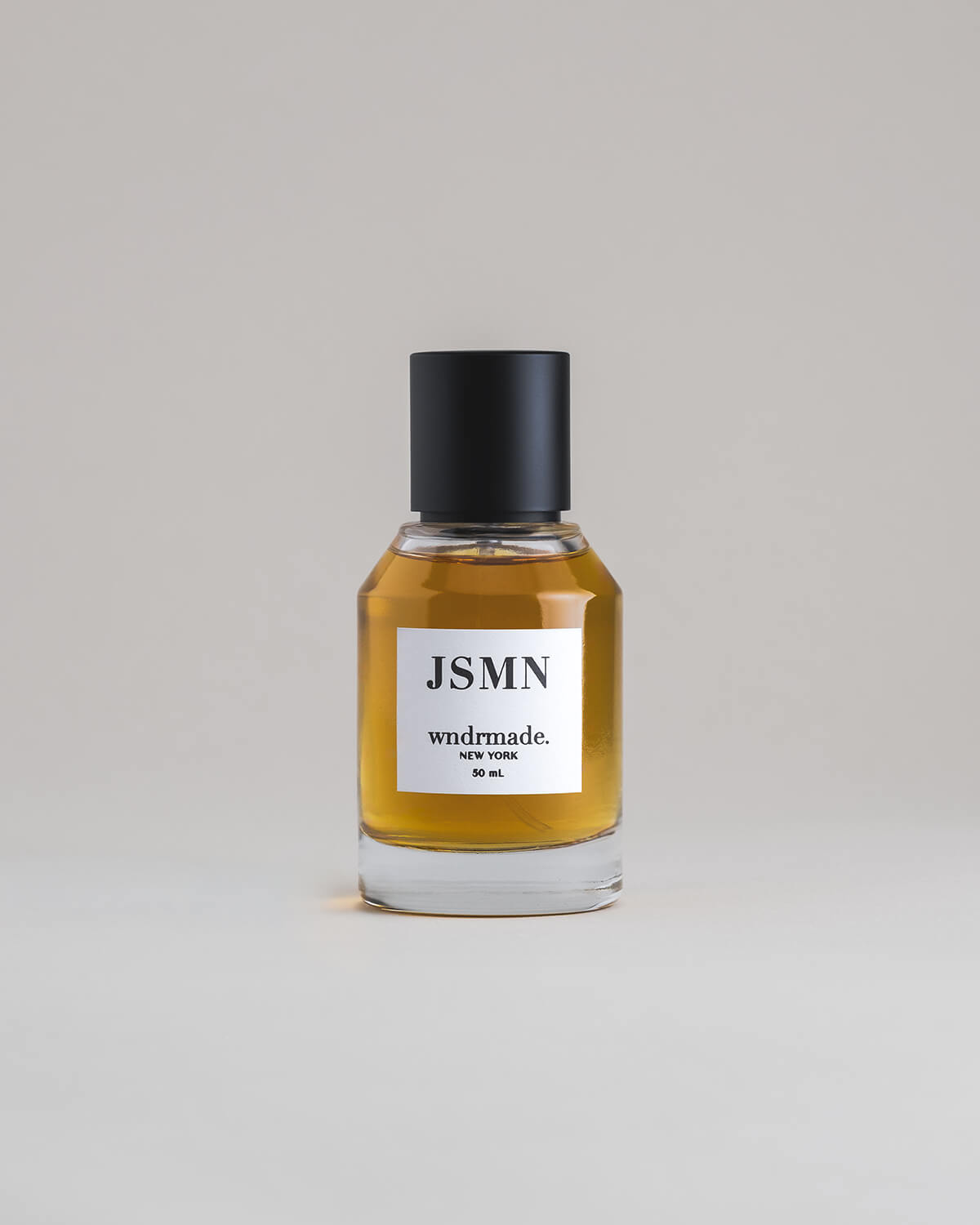 JSMN Jasmine 50 mL botanical perfume in a minimalist glass bottle with a matte black cap and a dark golden hue, set against a pale gray backdrop.