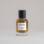 OKMS Oakmoss 50 mL botanical perfume in a minimalist glass bottle with a matte black cap and a dark golden hue, set against a pale gray backdrop.