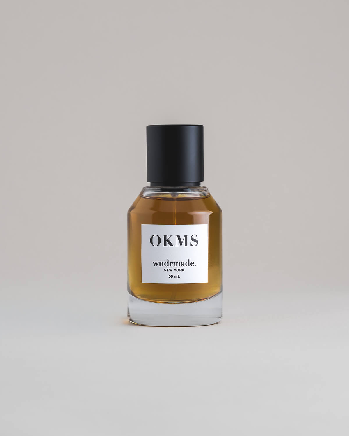 OKMS Oakmoss 50 mL botanical perfume in a minimalist glass bottle with a matte black cap and a dark golden hue, set against a pale gray backdrop.