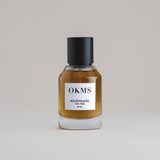 OKMS Oakmoss 50 mL botanical perfume in a minimalist glass bottle with a matte black cap and a dark golden hue, set against a pale gray backdrop.