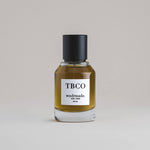 TBCO Tobacco 50 mL botanical perfume in a minimalist glass bottle with a matte black cap and a dark golden hue, set against a pale gray backdrop.