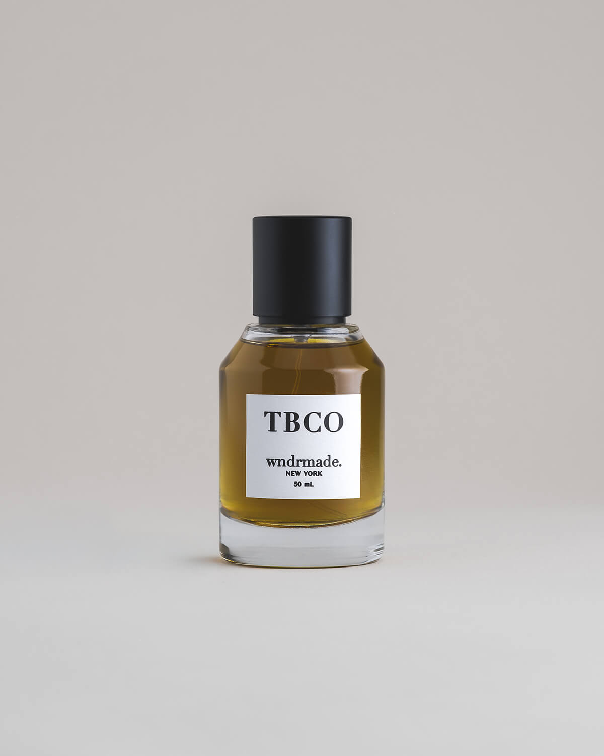 TBCO Tobacco 50 mL botanical perfume in a minimalist glass bottle with a matte black cap and a dark golden hue, set against a pale gray backdrop.